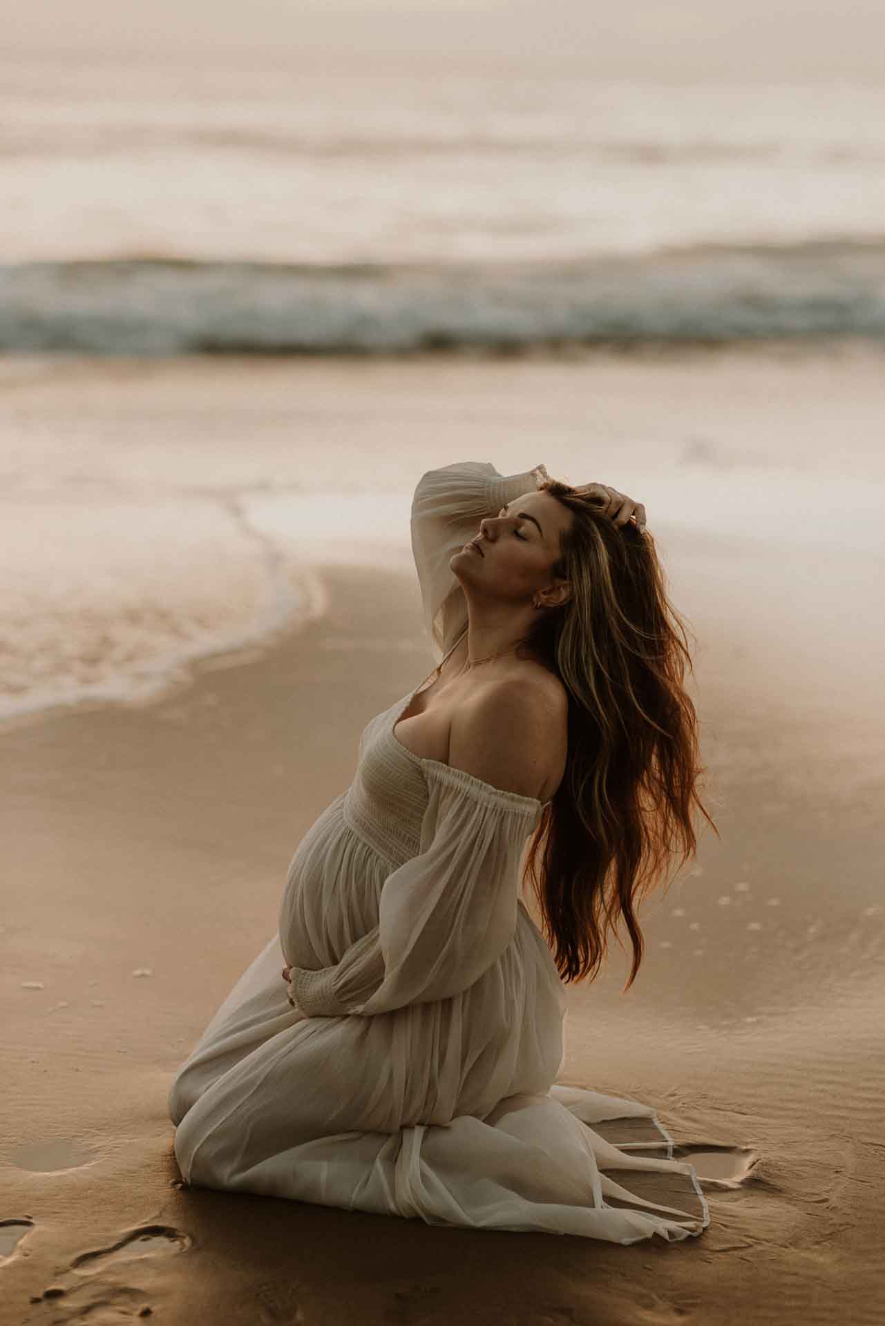 Pregnant woman sitting on the beach beside the ocean during a maternity session